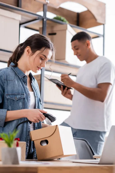 Vendeur numérisation boîte de carton près d'un ordinateur portable et confrère afro-américain flou dans la boutique en ligne — Photo de stock