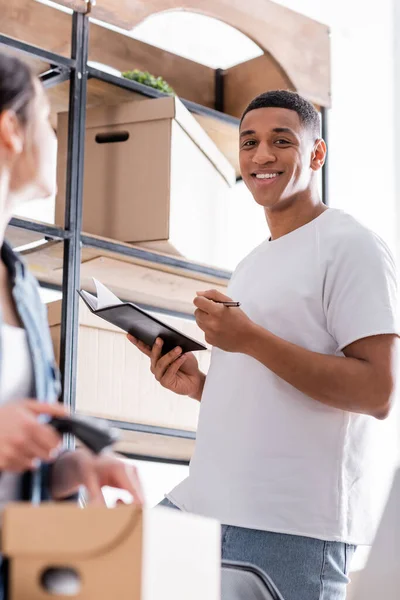 Alegre vendedor afroamericano celebración portátil cerca de colega borrosa con paquete en la tienda web en línea - foto de stock