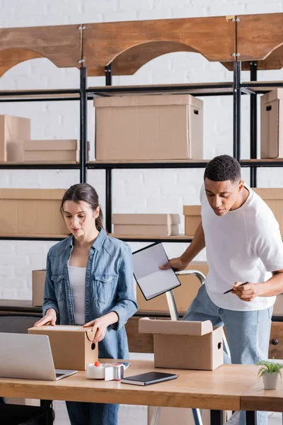 Vendeurs multiethniques travaillant avec des paquets près d'un ordinateur portable dans une boutique en ligne — Photo de stock