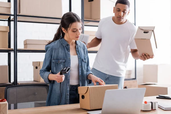 Vendedor escáner de retención cerca de caja de cartón, portátil y colega en la tienda web en línea - foto de stock