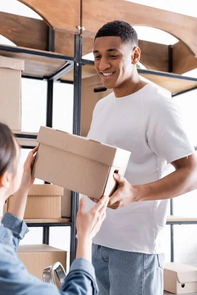 Lächelnder afrikanisch-amerikanischer Verkäufer hält Karton mit verschwommenem Kollegen in Online-Webshop — Stockfoto