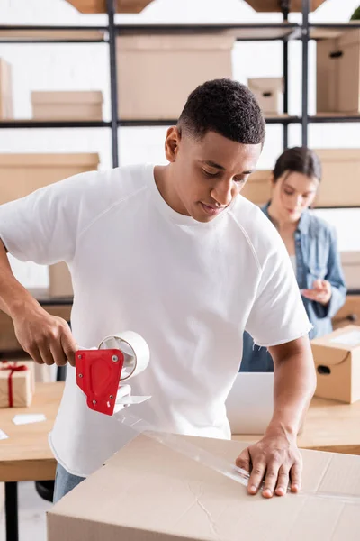 African american online web store owner packaging cardboard box with adhesive tape — Stock Photo