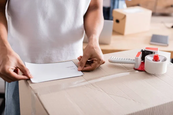 Cropped view of african american seller fastening shipping label on carton box near adhesive tape in online web store — Stock Photo