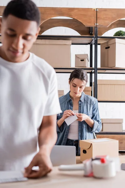 Joven vendedor sosteniendo etiqueta de envío cerca de caja de cartón y difuminado colega afroamericano en la tienda web en línea - foto de stock