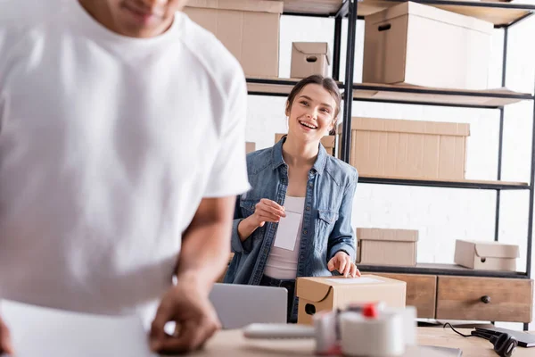 Vendedor sonriente sosteniendo etiqueta de envío cerca del paquete y difuminado colega afroamericano y tienda web en línea - foto de stock