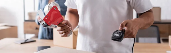 Cropped view of african american online web store owner holding scanner and adhesive tape near carton box, banner — Stock Photo