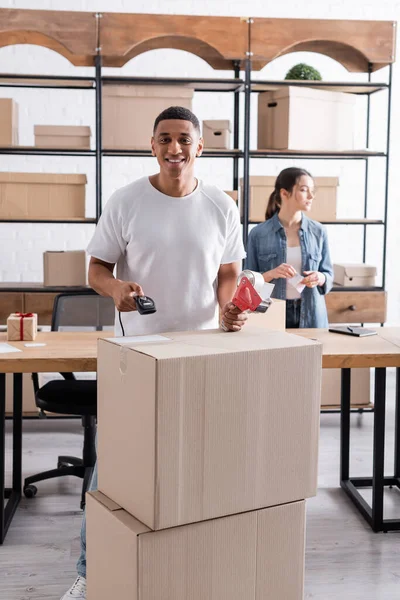 Propietario positivo de la tienda web afroamericana en línea que sostiene el escáner y la cinta adhesiva cerca de un colega borroso - foto de stock