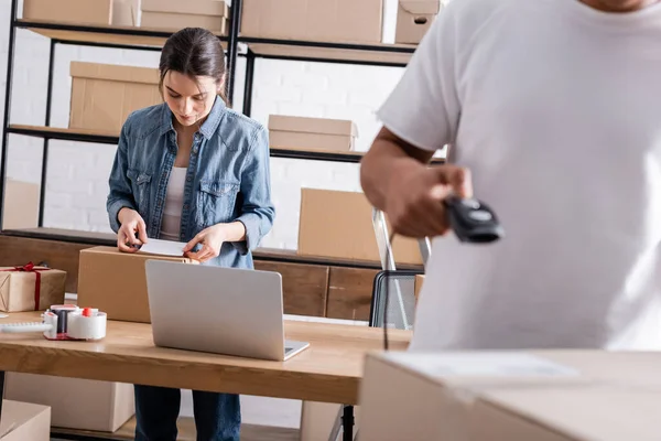 Seller fastening shipping label on cardboard box near laptop and african american colleague with scanner in online web store — Stock Photo