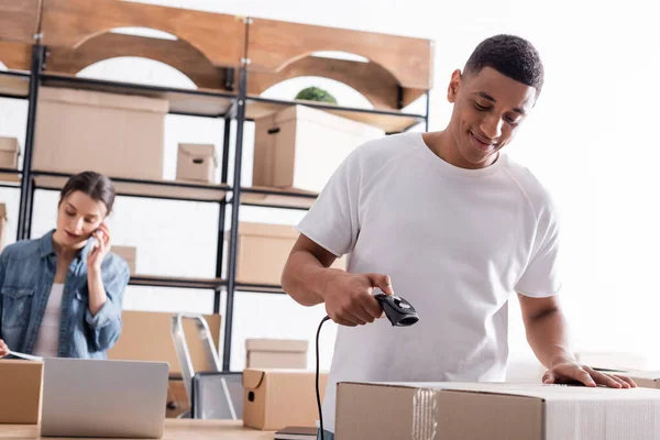 Sonriente vendedor afroamericano escaneo caja de cartón cerca de colega borrosa en la tienda web en línea — Stock Photo