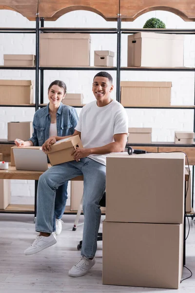 Propietarios positivos multiétnicos de tiendas web en línea mirando la cámara cerca de la computadora portátil y paquetes en rack - foto de stock