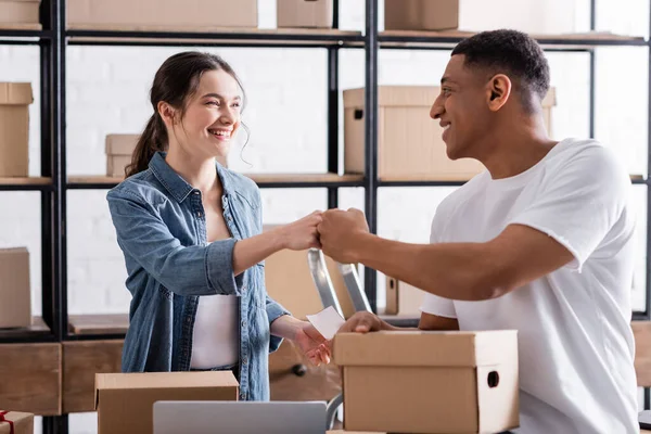 Positive multiethnic sellers doing fists bump near packages in online web store — Stock Photo