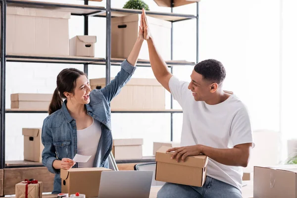 Alegre multiétnicos vendedores dando alta cinco cerca de cajas de cartón y portátil en la tienda en línea web - foto de stock