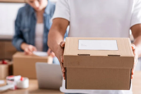 Cropped view of african american seller holding box with shipping label near blurred colleague in online web store — Stock Photo