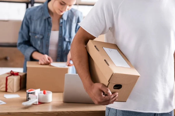 Vendedor afroamericano sosteniendo caja de cartón cerca de colega borroso que trabaja en la tienda web en línea - foto de stock