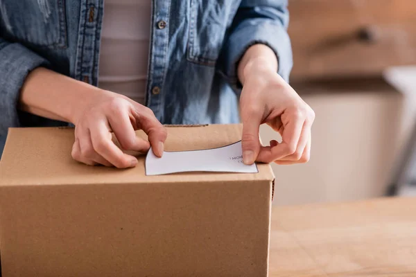 Cropped view of seller fastening shipping label on cardboard box in online web store — Stock Photo