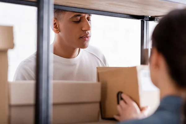 African american seller standing near packages and blurred colleague in online web store — Stock Photo