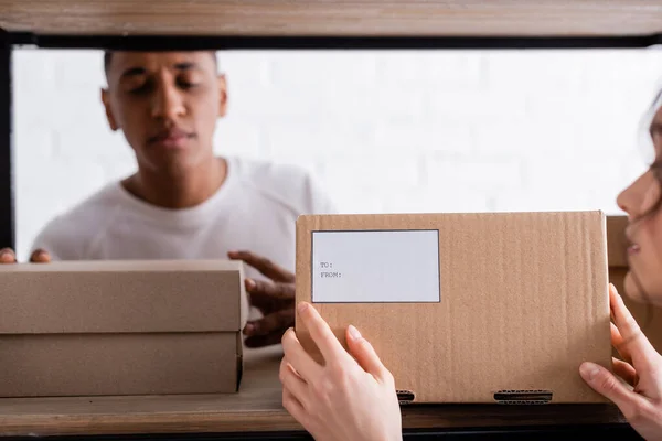 Vendedor borroso sosteniendo caja de cartón con etiqueta de envío cerca borrosa colega afroamericano en la tienda web en línea - foto de stock