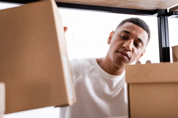 African american seller looking at blurred packages on rack in online web store — Stock Photo