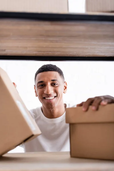 Sonriente vendedor afroamericano mirando a la cámara cerca de cajas de cartón borrosas en rack en la tienda web en línea - foto de stock