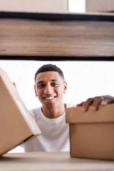 Sorrindo vendedor americano africano olhando para a câmera perto de caixas de papelão borradas no rack na loja online — Fotografia de Stock