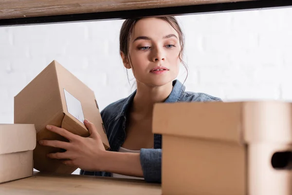 Joven vendedor sosteniendo paquete cerca de caja de cartón borrosa en rack en la tienda web en línea - foto de stock