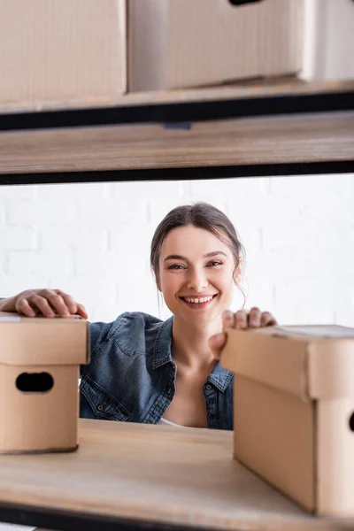 Venditore sorridente guardando la fotocamera vicino scatole di cartone sfocate su rack nel negozio online web — Foto stock