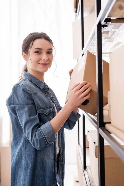 Sorrindo vendedor segurando caixa e olhando para a câmera perto rack na loja online — Fotografia de Stock