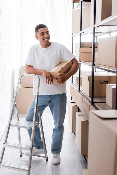 Le vendeur afro-américain positif tenant la boîte de carton près du rack et de l'échelle dans la boutique en ligne — Photo de stock