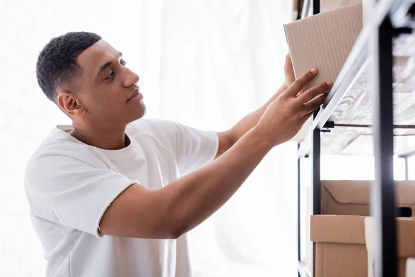 Afrikanisch-amerikanischer Verkäufer hält Karton in der Nähe von Regal in Online-Webshop — Stockfoto