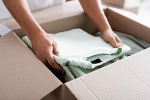 Vista recortada del vendedor afroamericano poniendo ropa en caja de cartón en tienda web en línea - foto de stock