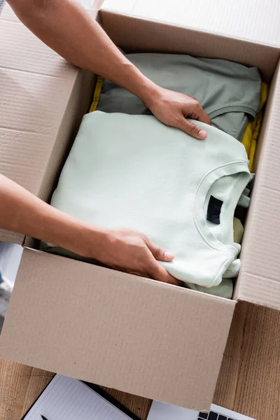 Cropped view of african american owner putting clothes in carton box in online web store — Stock Photo