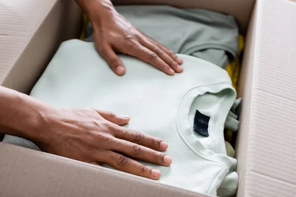 Cropped view of african american seller putting clothes in package in online web store — Stock Photo