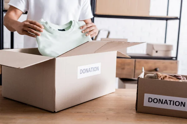 Vista recortada del vendedor afroamericano poniendo ropa en caja con letras de donación en la mesa - foto de stock