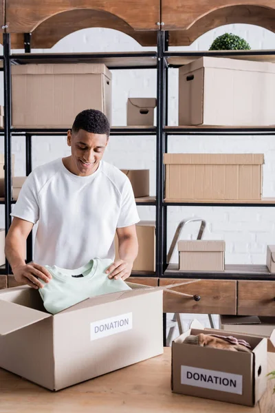 Smiling african american owner putting clothes in box with donation lettering in online web store — Stock Photo