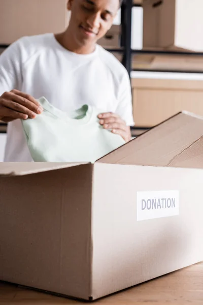 Caja con letras de donación cerca del borroso propietario afroamericano con ropa en la tienda web en línea - foto de stock