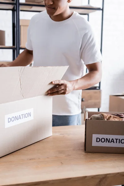 Cropped view of african american owner of online web store holding box with donation lettering on table — Stock Photo