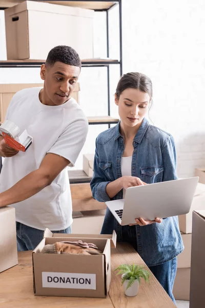 Seller holding laptop near african american colleague with adhesive tape and boxes with donation lettering in online web store — Stock Photo