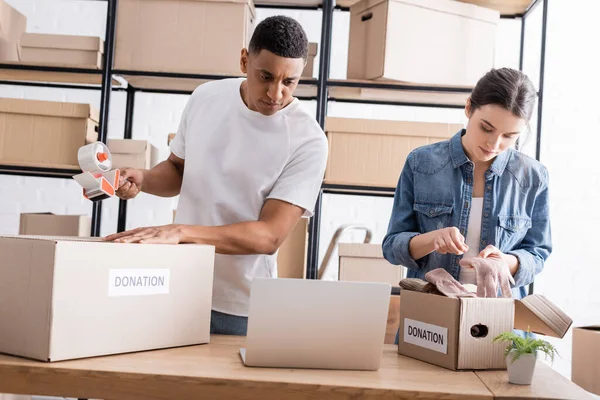Interracial sellers packaging boxes with donation lettering near laptop in online web store — Stock Photo