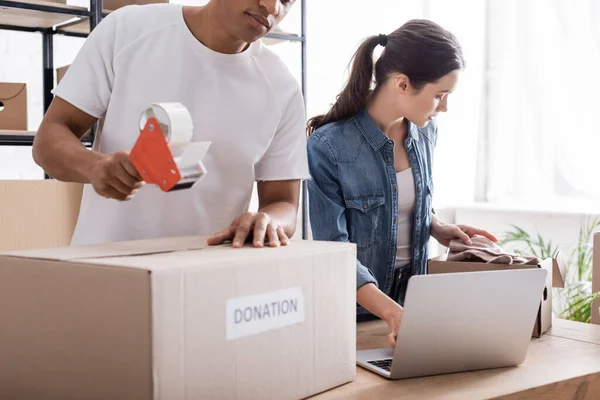 Multiethnic sellers packaging boxes with donation lettering near laptop in online web store — Stock Photo