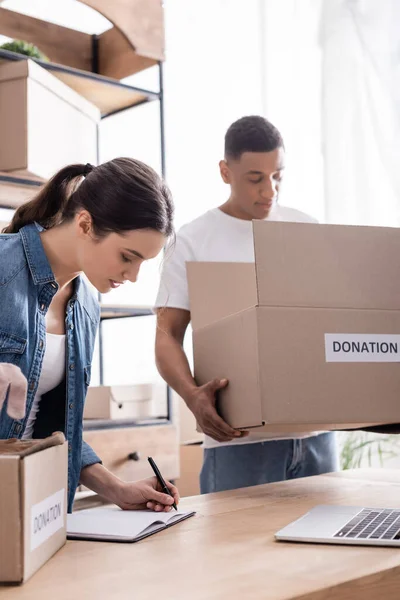 Joven vendedor escribiendo en un cuaderno cerca de la computadora portátil y borrosa colega afroamericano con caja de donaciones en la tienda web en línea - foto de stock