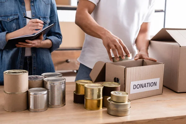 Vista recortada de vendedores multiétnicos escribiendo en cuaderno y caja de donación de envases cerca de alimentos enlatados en la mesa - foto de stock