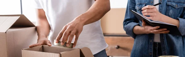 Vista recortada del vendedor afroamericano poniendo comida enlatada en caja cerca de colega con cuaderno en la tienda web en línea, pancarta - foto de stock