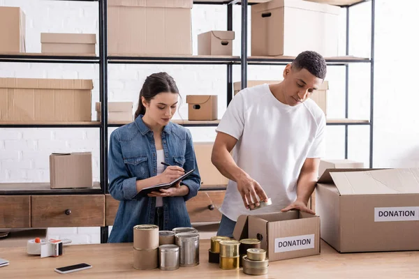 Multiethnic sellers packaging box with donation lettering near canned food in online web store — Stock Photo