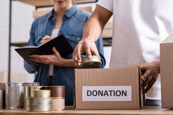 Vista recortada del vendedor afroamericano sosteniendo comida enlatada cerca de la caja con letras de donación y colega borroso en la tienda web en línea - foto de stock