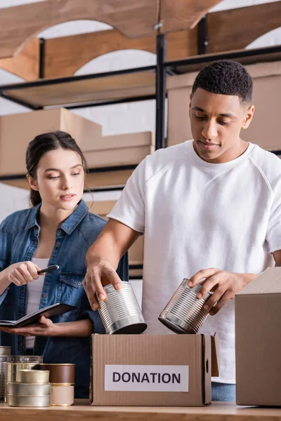 Proprietários de lojas online multiétnicas empacotando alimentos enlatados em caixa com letras de doação na mesa — Fotografia de Stock