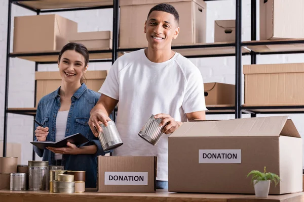 Lächelnde multiethnische Online-Shop-Betreiber halten Konserven in der Nähe von Schachteln mit Spendenaufdruck auf dem Tisch — Stockfoto