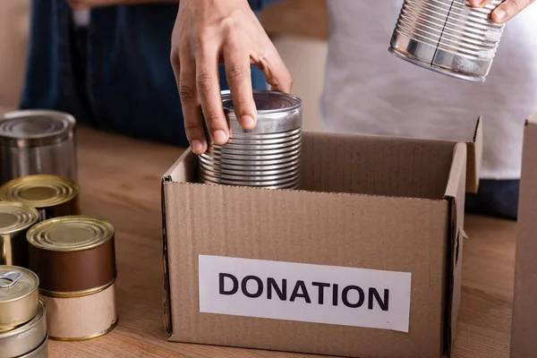 Cropped view of african american seller putting canned food n carton box with donation lettering in online web store — Stock Photo