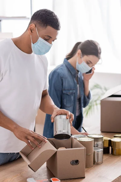 Africano americano vendedor em máscara médica colocando alimentos enlatados em caixa de papelão na loja online — Fotografia de Stock