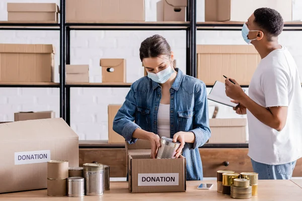 Vendeurs multiethniques dans des masques médicaux mettant de la nourriture en conserve dans une boîte avec des lettres de dons dans une boutique en ligne — Photo de stock