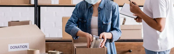 Vista recortada del vendedor poniendo comida enlatada en la caja de donación cerca de un colega afroamericano en la tienda web en línea, pancarta - foto de stock